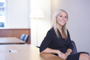Lady with blonde hair smiling at camera , seated with her elbow on desk.